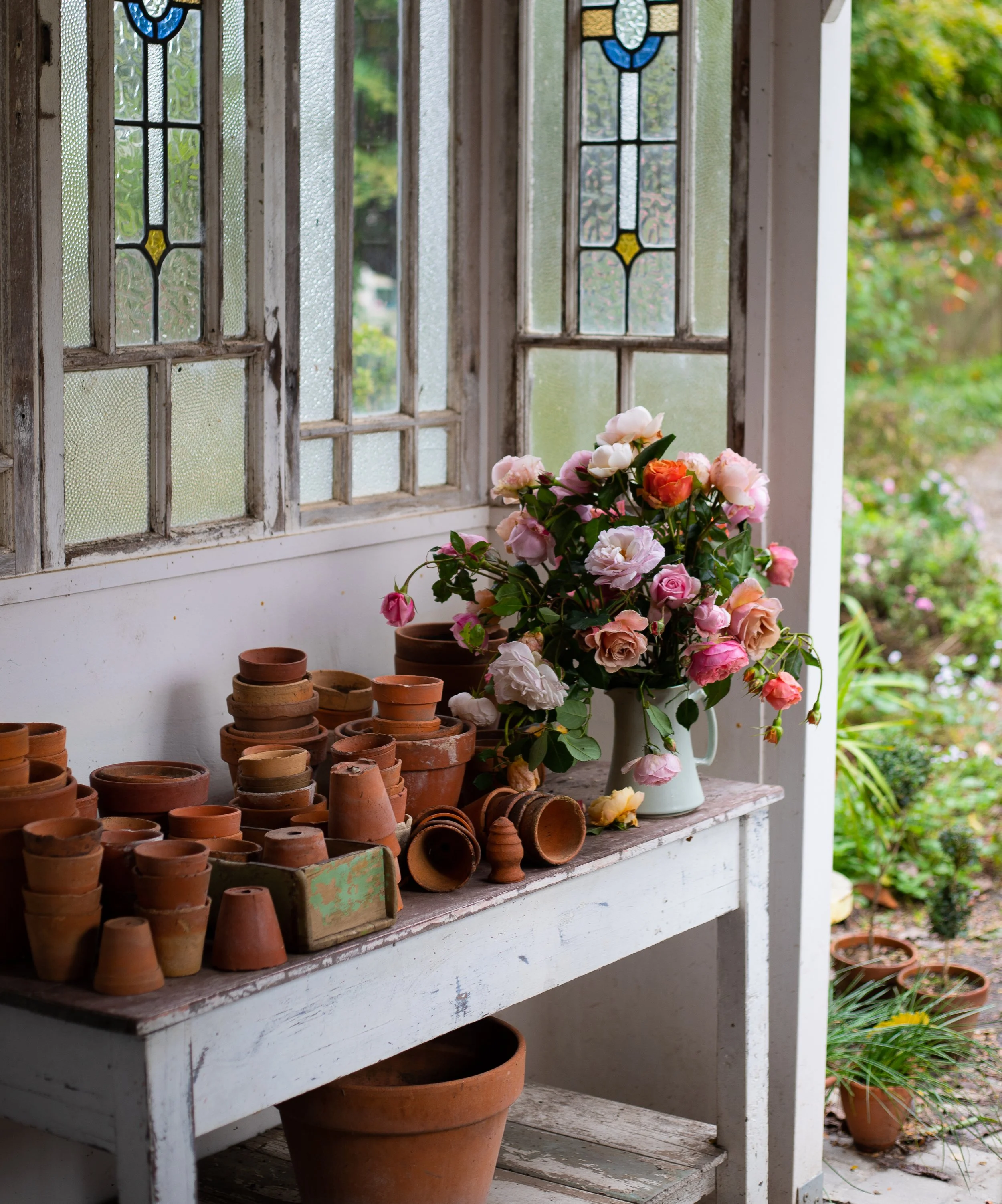 A windowsill with a large bouquet of pink and white roses in a white pitcher, surrounded by empty terracotta pots, inside a rustic room with stained glass windows.