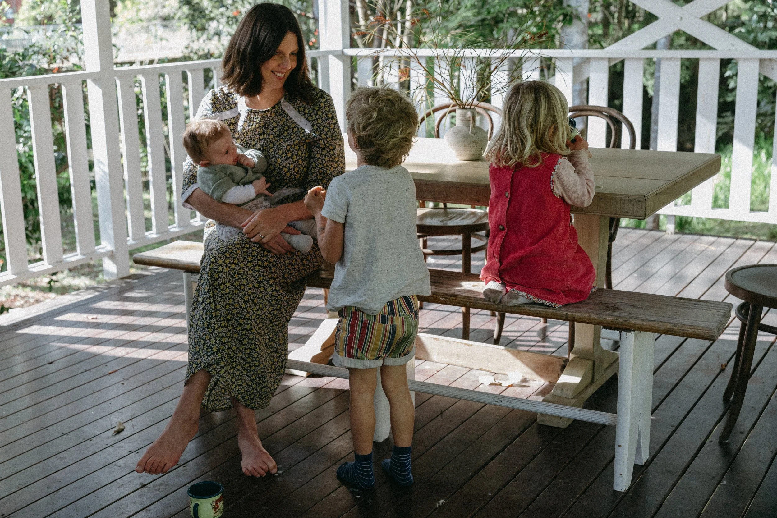 A woman with shoulder-length dark hair, wearing a floral dress, sitting at a porch table, holding a baby, and smiling at three children who are standing and sitting nearby.
