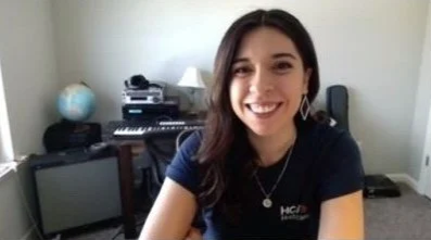 Young woman with dark hair smiling, sitting in a room with a keyboard, speaker, and desk in the background.