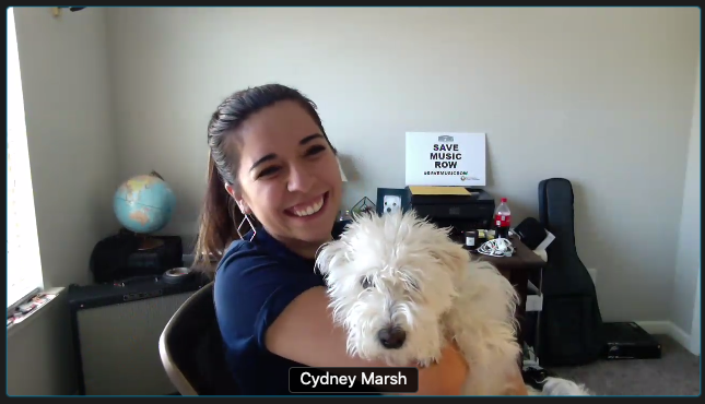 Smiling woman named Cydney Marsh holding a fluffy white dog in a home office with a globe, books, and a sign that reads 'SAVE MUSIC ROW' in the background.