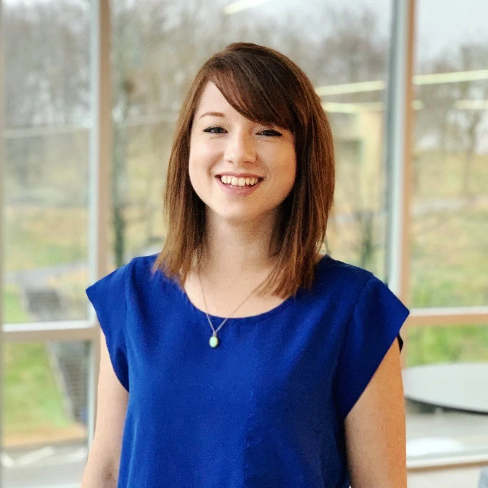 A young woman with shoulder-length brown hair, smiling, wearing a blue top and a necklace with a light-colored pendant, standing indoors near large windows.