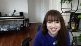 Young woman with brown hair and bangs smiling in a room with hardwood floors, a bed, and a black metal shelf.