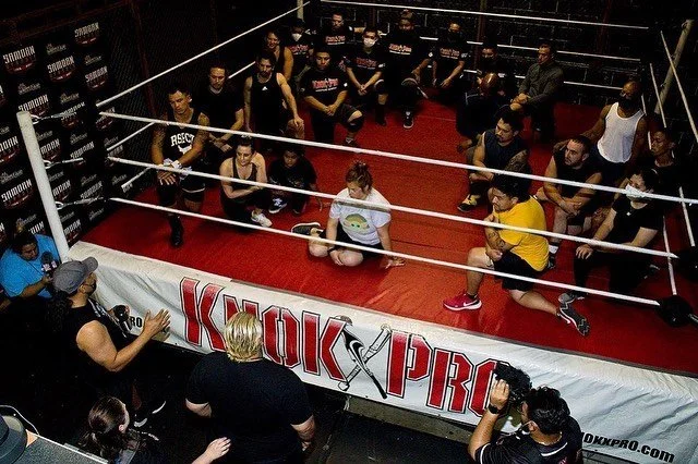 People seated on the floor inside a wrestling ring during a class or seminar, with instructors and audience members around the ring.