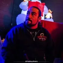 A man sitting in a dimly lit room with Christmas decorations, including a snowman and a Christmas tree, in the background.