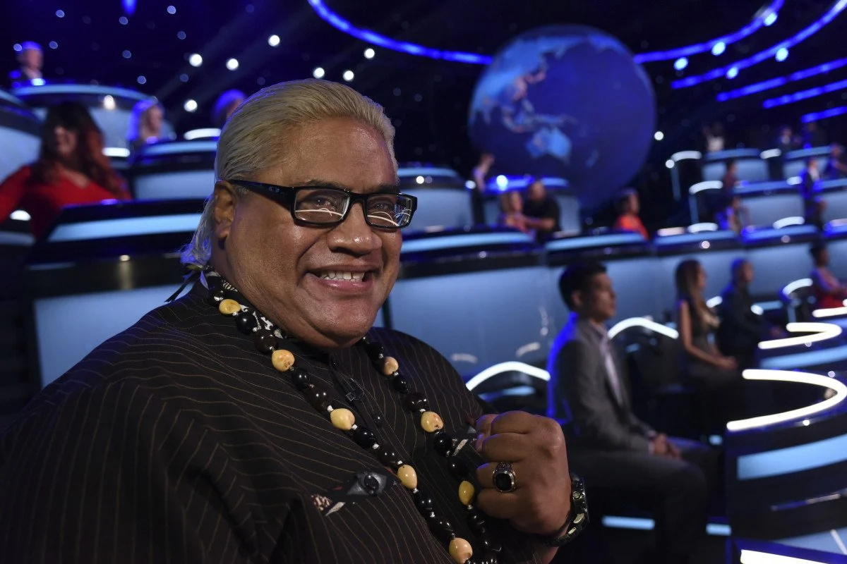 Smiling man with glasses and long gray hair, wearing a beaded necklace, in a brightly lit studio with a large globe and多people seated in a tiered arrangement.