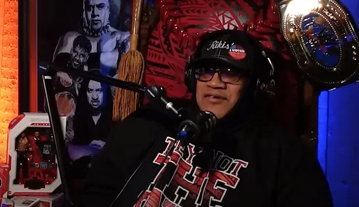 A man wearing glasses, a black hoodie, and a cap that reads 'I VOTED' is sitting in front of a microphone in a recording studio. The background features black and white artwork, a red patterned wall, and a large gold championship belt hanging on the wall.