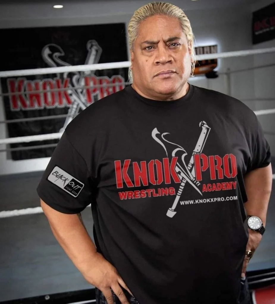 A man with gray hair is standing in a wrestling gym, wearing a black t-shirt with 'Knock Pro Wrestling Academy' printed on it and a logo of a crutch, knife, and syringe. The background shows a wrestling ring.