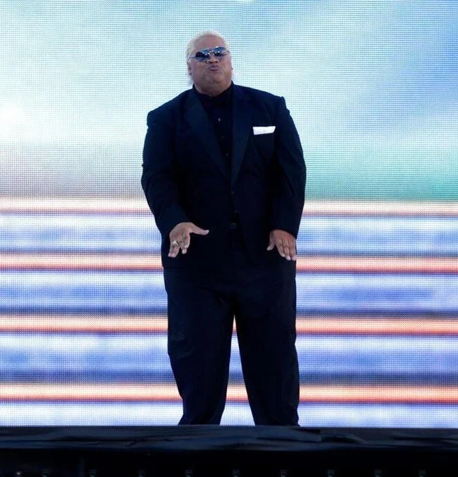 Man with white hair and sunglasses wearing a black suit and black shirt standing on stage with a colorful digital background.