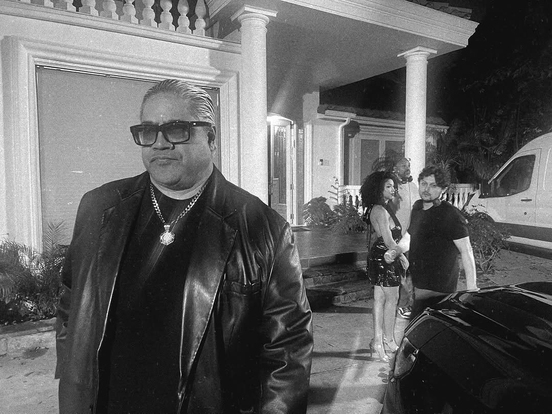 A black-and-white photo of four people outside a house at night. The man in the foreground is wearing sunglasses, a leather jacket, and chains. The three people in the background are holding hands, with two women dressed in patterned dresses and a man in casual black clothing.