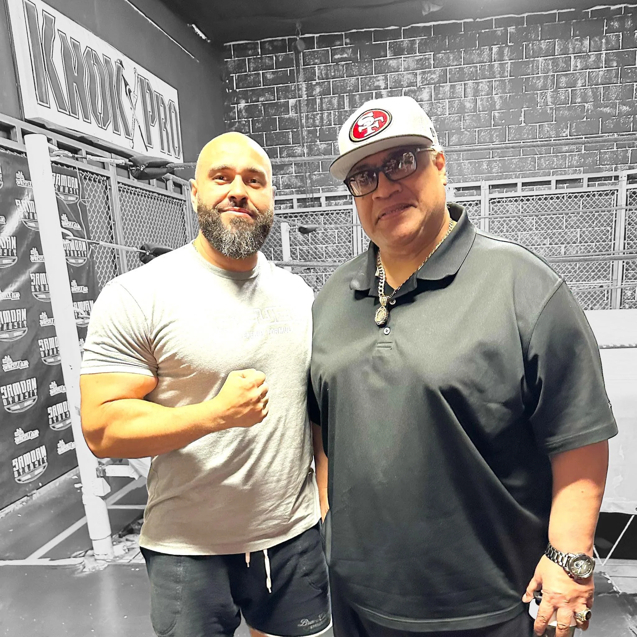 Two men standing inside a boxing gym, with the man on the left flexing his arm and the man on the right wearing a cap, sunglasses, and jewelry, both looking at the camera.