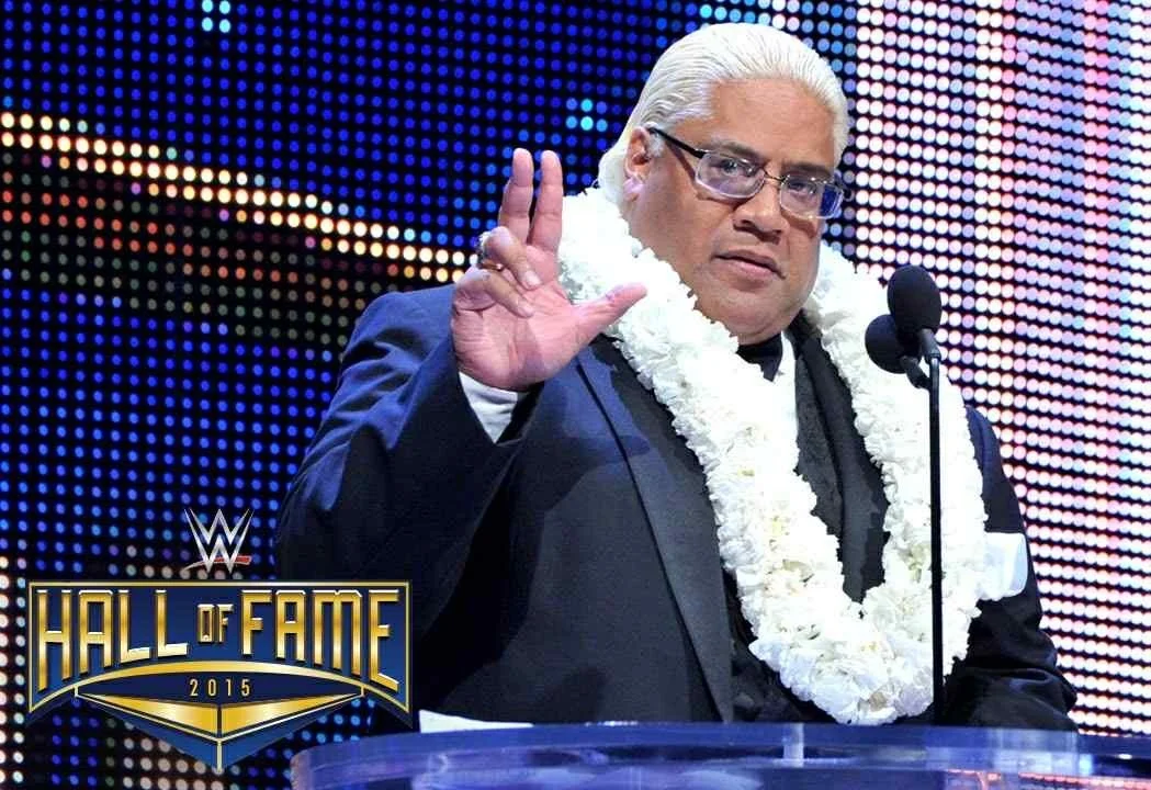 A man with platinum blonde hair in a suit, wearing glasses and a lei, making a gesture with his hand on a stage with a WWE Hall of Fame logo in 2015.