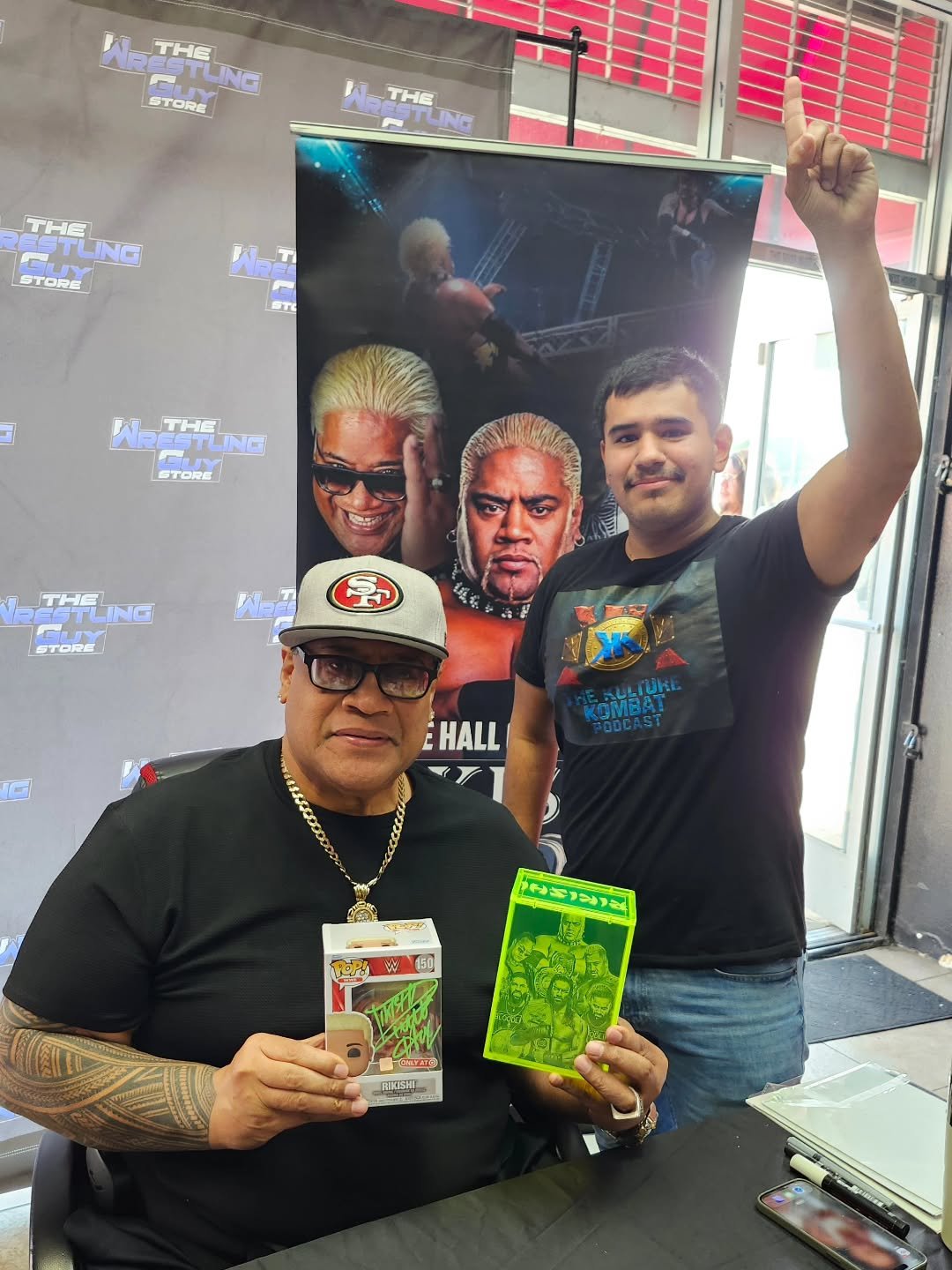 Two men, one older and one younger, poses at a wrestling event. The older man holds a Funko Pop figure and a neon green collectible card, both featuring wrestling characters. The background features a promotional poster and a gray backdrop with the logo "The Wrestling Guy Store." The younger man is making a gesture with his right hand represented by his index finger raised, and is wearing a black T-shirt with a ``Kano'' design from Mortal Kombat.