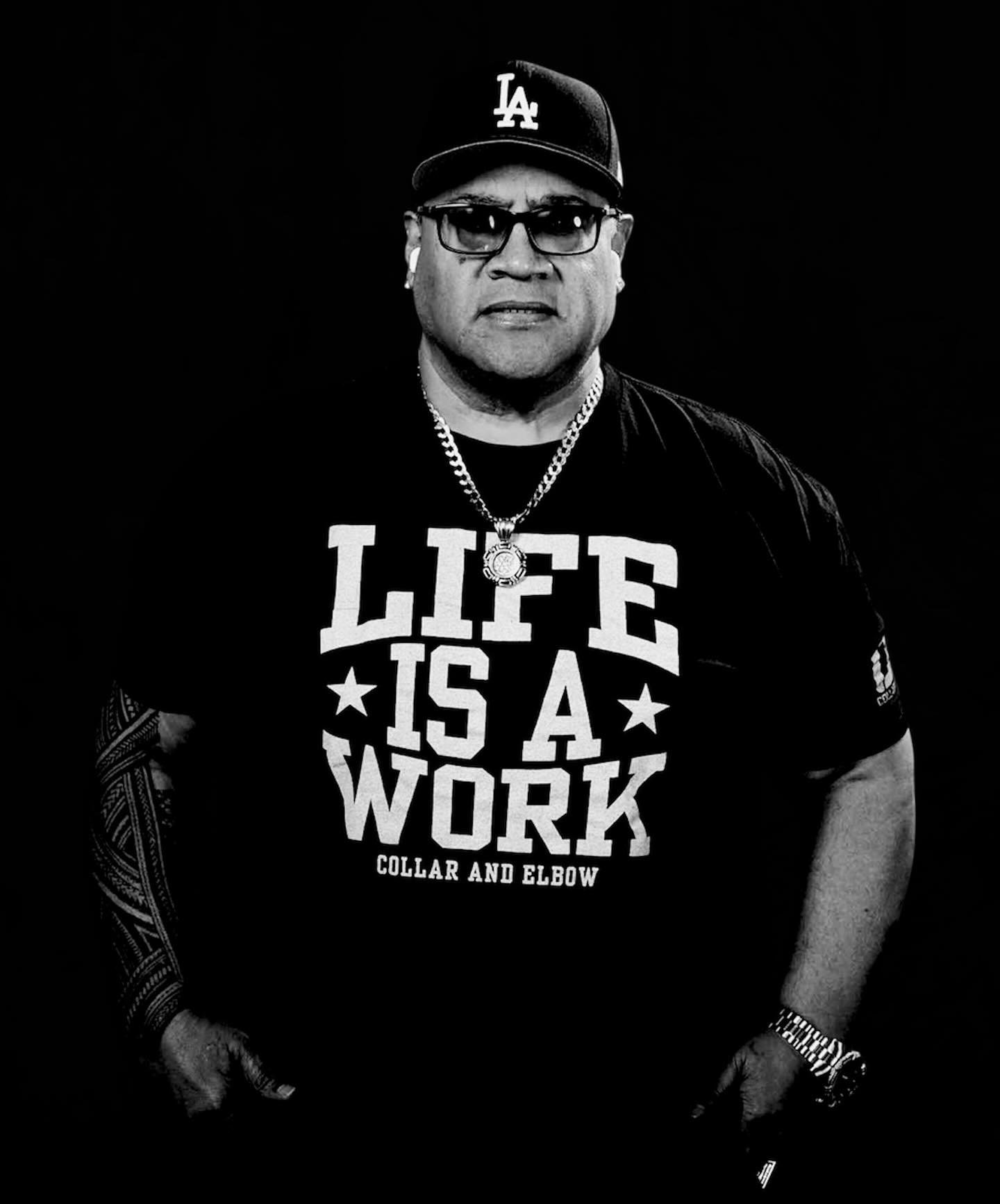 Man wearing a baseball cap, sunglasses, and a black T-shirt that says 'Life is a work, collar and elbow'. He has tattoos, jewelry, and earbuds.