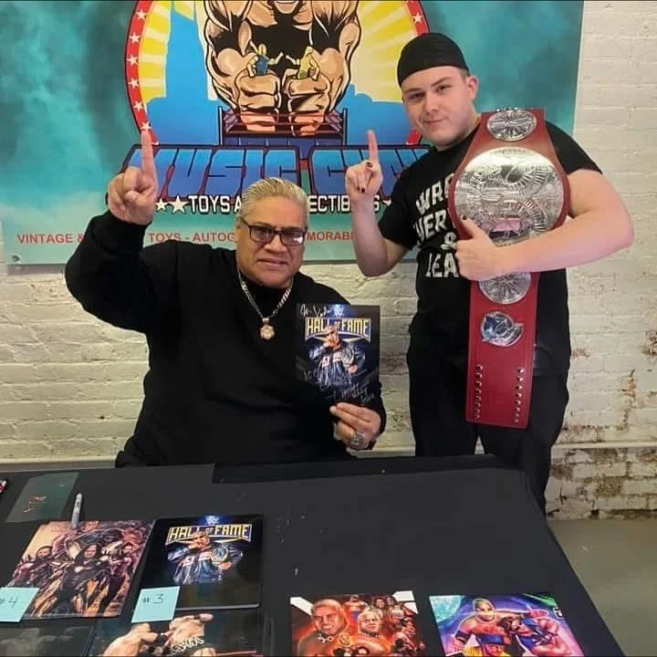 A young man wearing a black T-shirt and cap, holding a championship belt, stands next to an older man with glasses, holding a Hall of Fame magazine, at a signing event. They are pointing upward with their index fingers, in front of a wrestling-themed poster. The table has various comic and wrestling art prints.