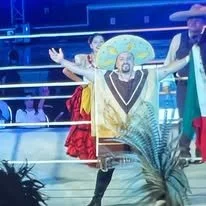 A performer on stage with arms outstretched, dressed in traditional attire, with two people in the background and a tropical plant in the foreground.