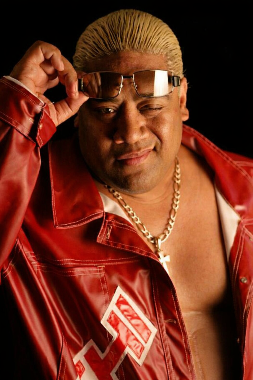 A man with bleached blonde hair, wearing a red leather jacket with white lettering, a chunky gold chain with a cross pendant, and sunglasses that he is lifting with his right hand. He is smiling and winking at the camera against a black background.