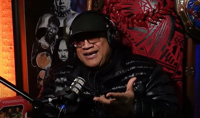 A man in a black leather jacket and black cap speaking into a microphone, with headphones on, in a recording studio.