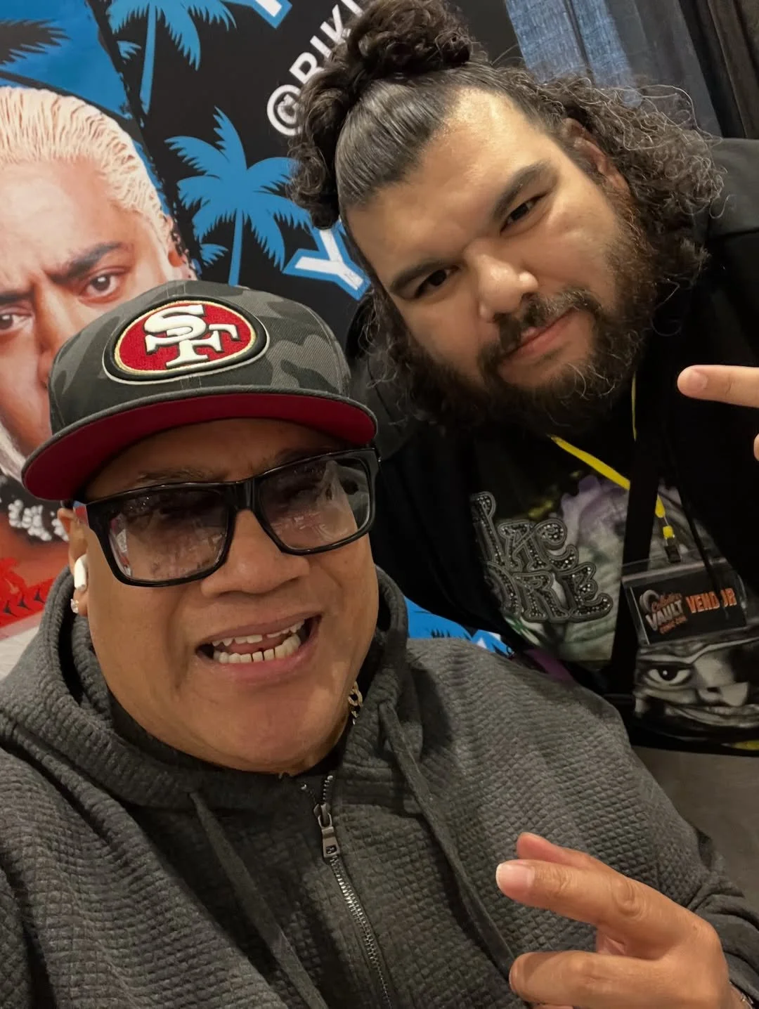 Two men taking a selfie, one wearing a San Francisco 49ers cap and glasses, the other with long curly hair and a beard, making a peace sign. Behind them are posters, including a large face of a man with white hair and a black background with palm trees and the word 'Venom'.