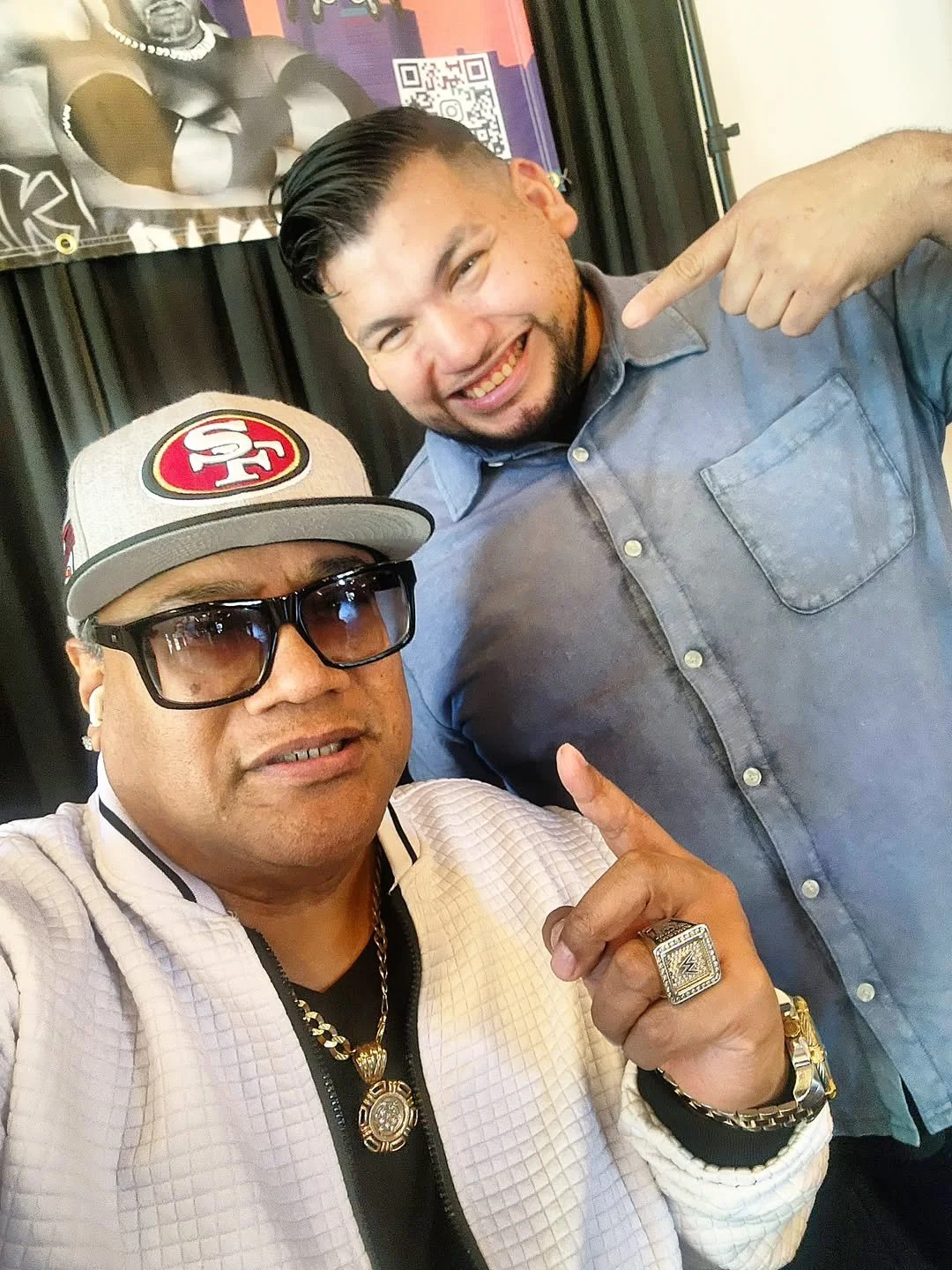 Two men taking a selfie together. The man in front is wearing sunglasses, a baseball cap with an SF logo, and jewelry. The man behind is smiling and pointing at the man in front. They appear to be at an indoor event with a backdrop featuring promotional material and a QR code.