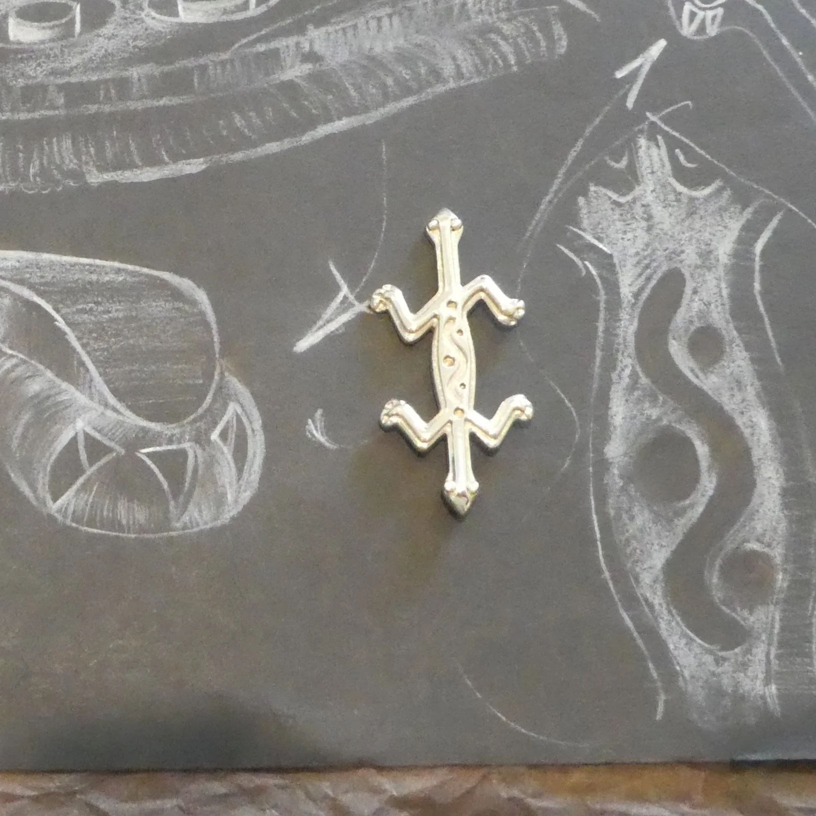 A metallic Cross pendant placed on a black surface with white chalk sketches of a snake, a fish, a skull, and a spiral design drawn around it.
