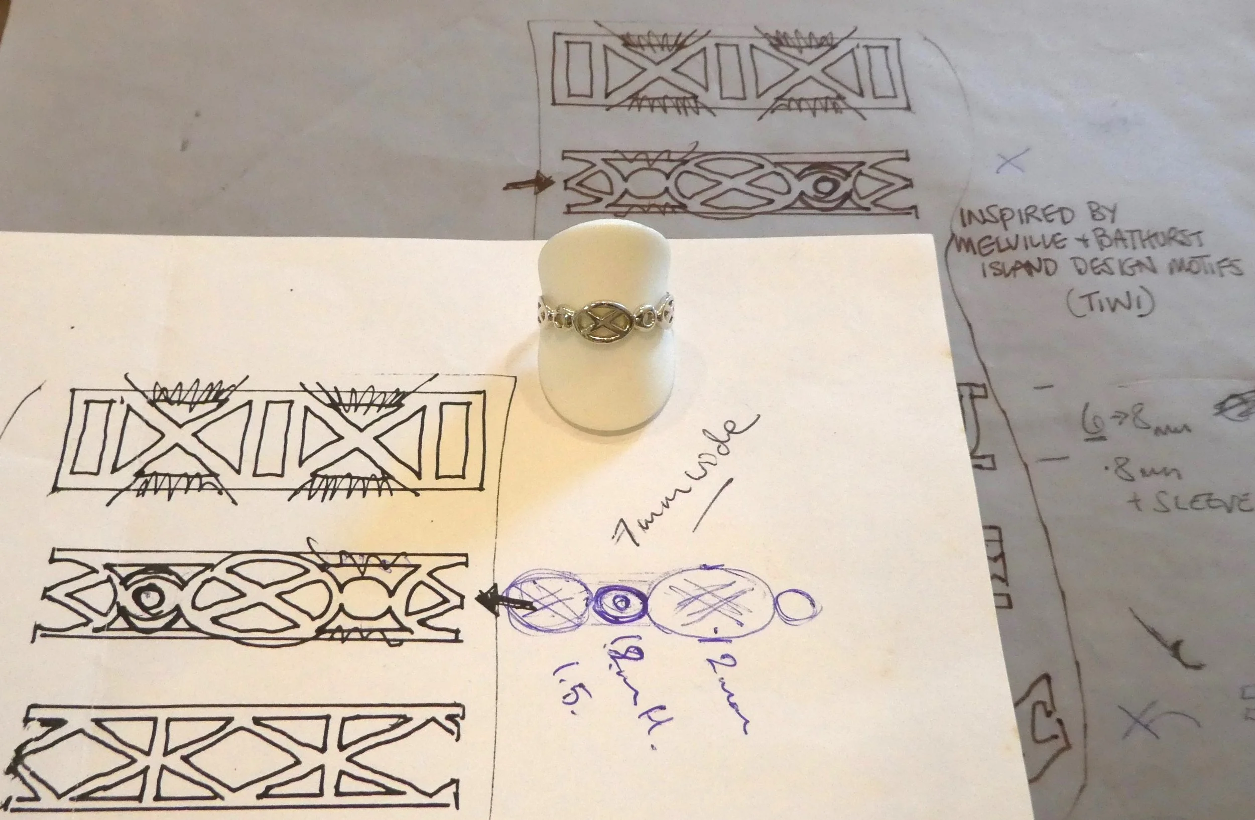 A ring with indigenous island-inspired motifs is displayed on a beige model finger, with hand-drawn sketches and notes about the design behind it, inspired by Melville and Bathurst island motifs.