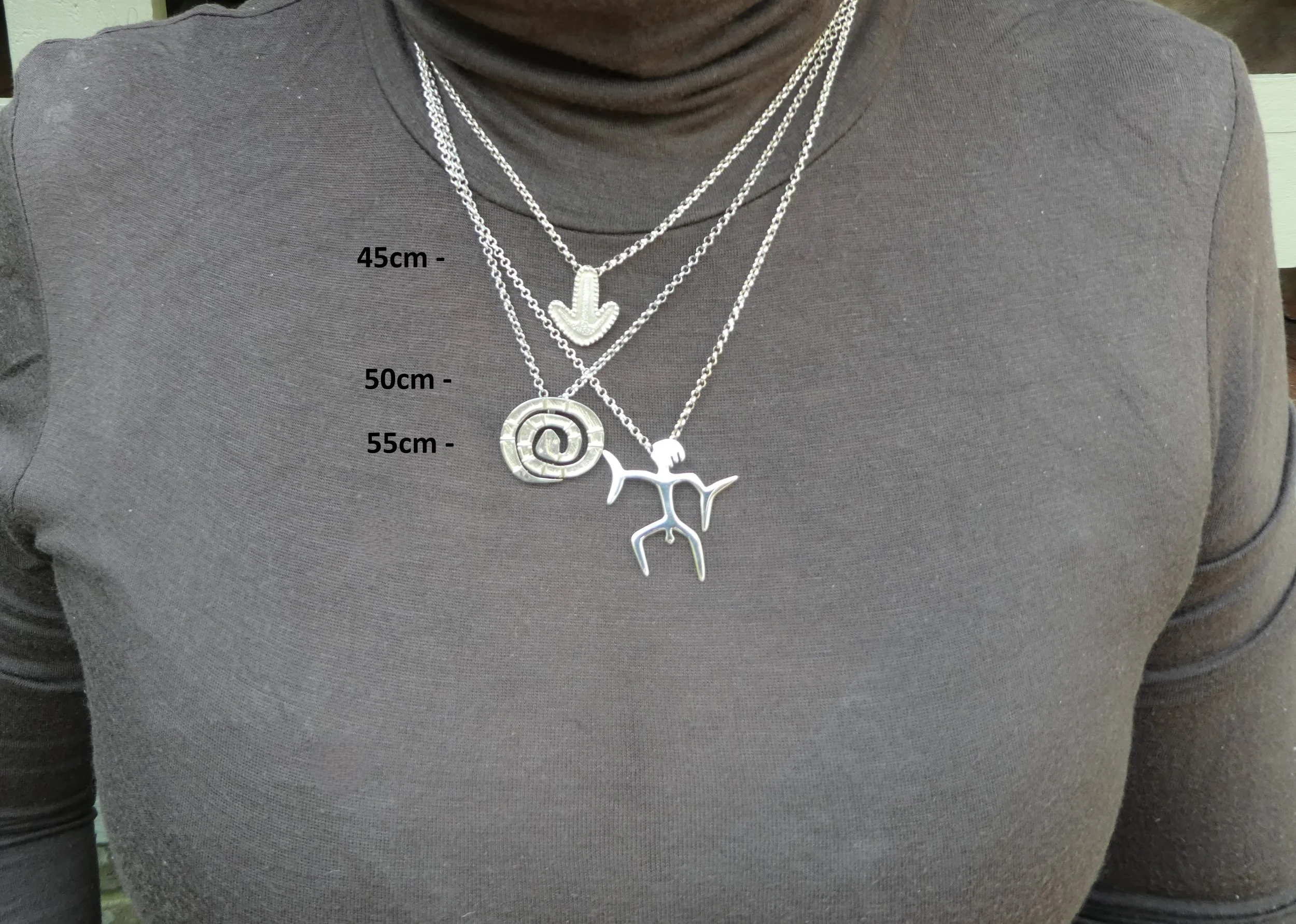 Close-up of a person wearing a dark gray turtleneck shirt with three layered silver necklaces. The necklaces have different charms: a downward arrow, a spiral, and a stylized human figure with outstretched arms and legs. Measurements in centimeters indicate the necklace lengths: 45cm, 50cm, and 55cm.