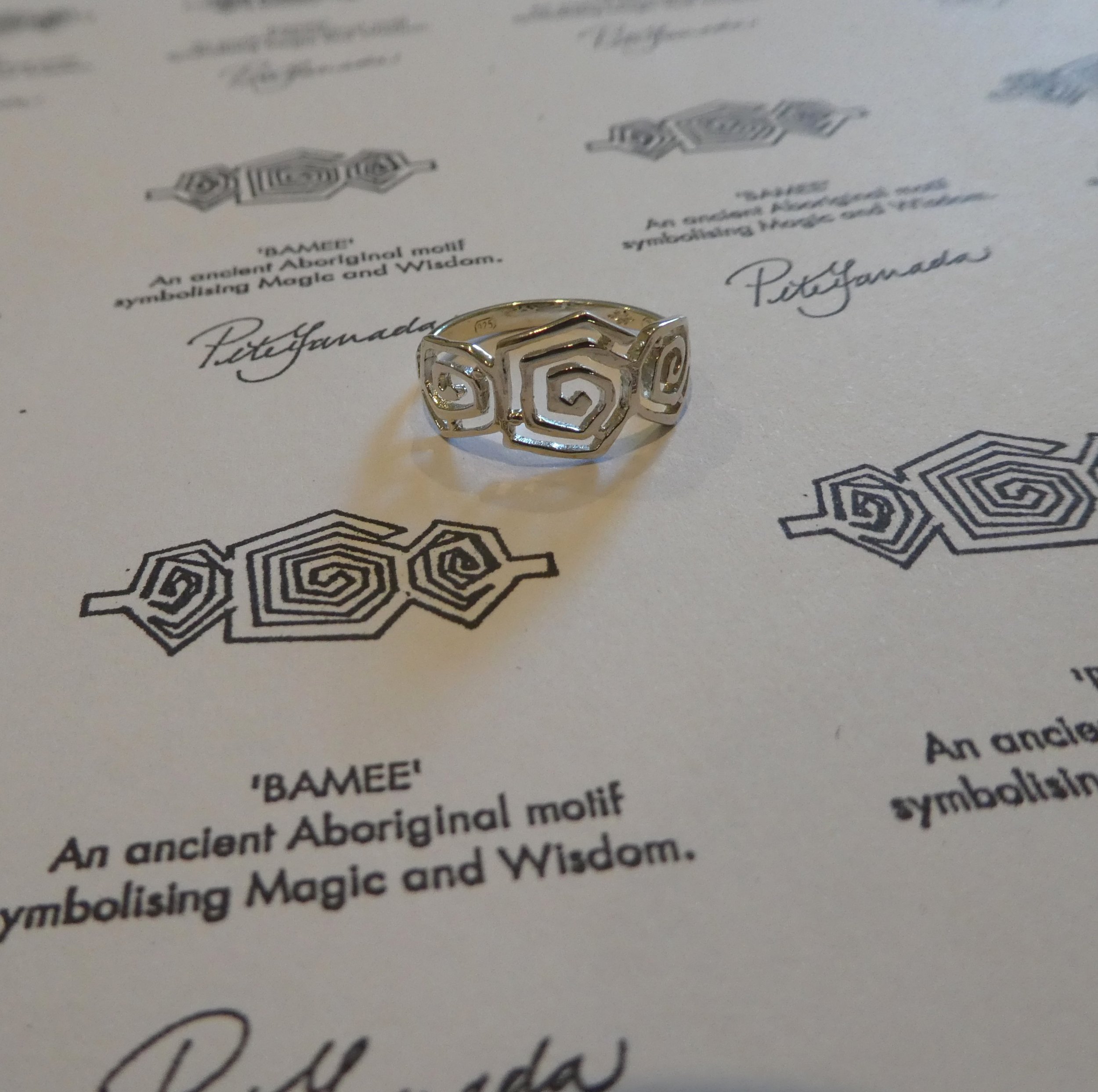 A silver spiral ring placed on a sheet of paper with printed Aboriginal motifs and signatures.