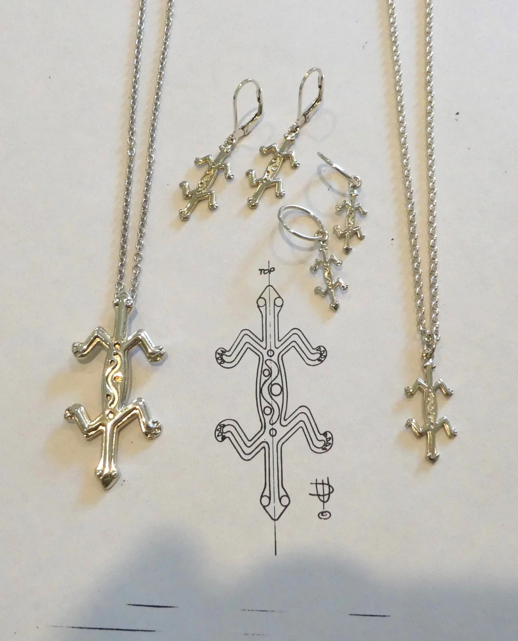 Silver jewelry set including necklaces, earrings, a ring, and a bracelet, all featuring a gecko design, placed on a white surface with a schematic drawing of the gecko jewelry design.