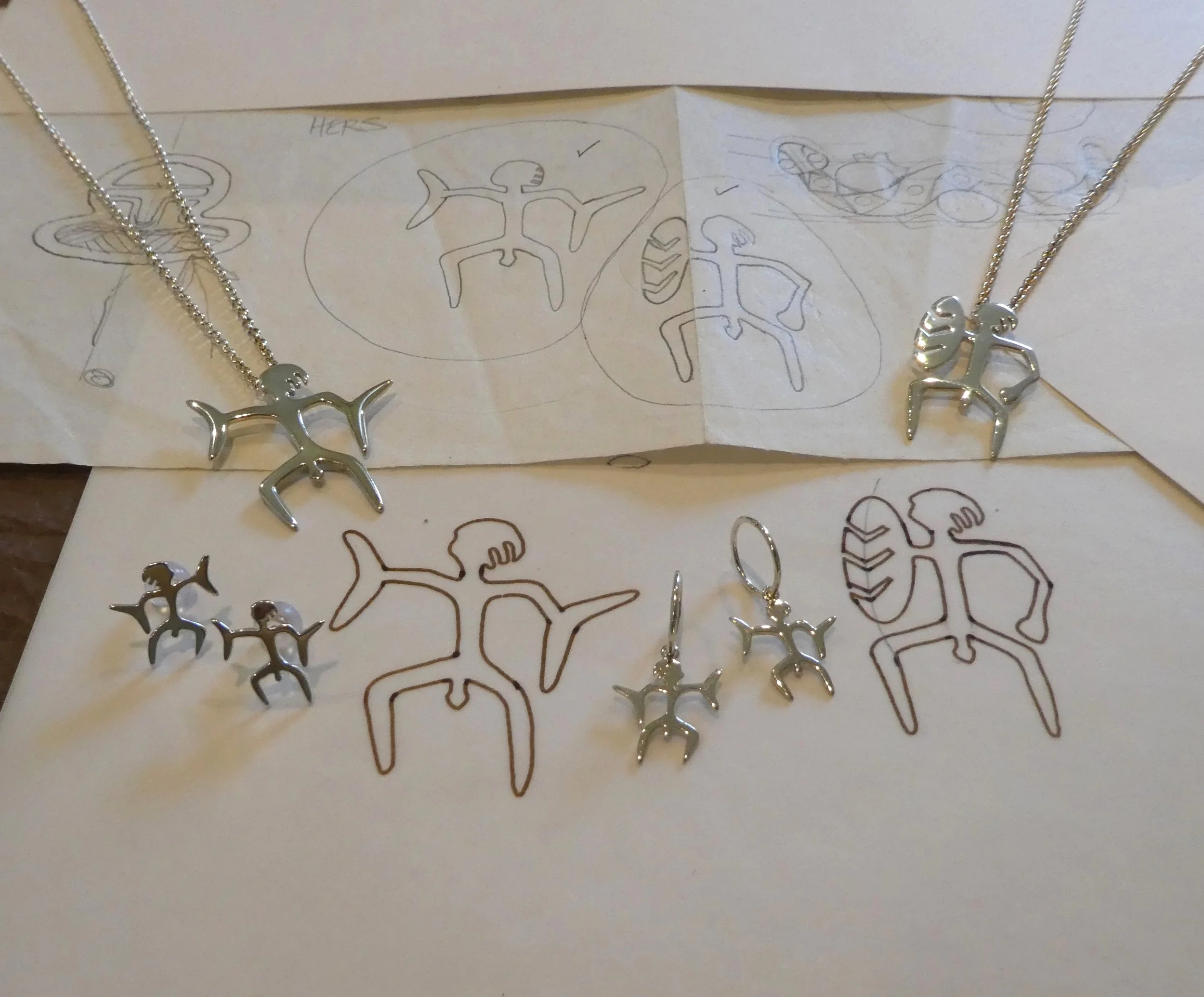 Silver jewelry set including two necklaces, a pair of earrings, and a pair of cufflinks, all featuring a human-like figure with outstretched arms and a fish on the head, placed on sketches of the jewelry design.