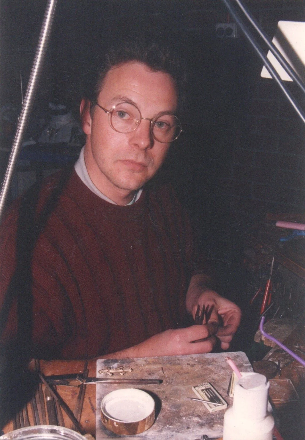 A man with glasses working at a cluttered wooden desk, surrounded by tools and electronic components.