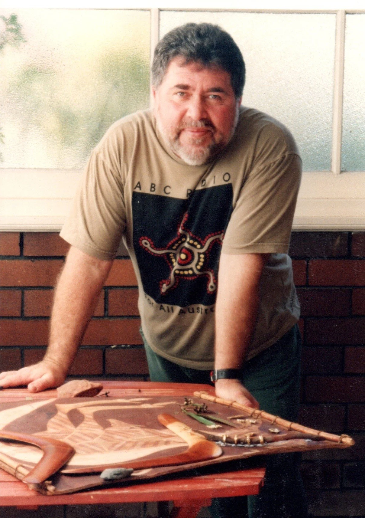 A man with gray hair and beard, wearing a beige T-shirt with a graphic design, leaning on a table with a wooden boomerang and some small objects, in front of a brick wall and window.