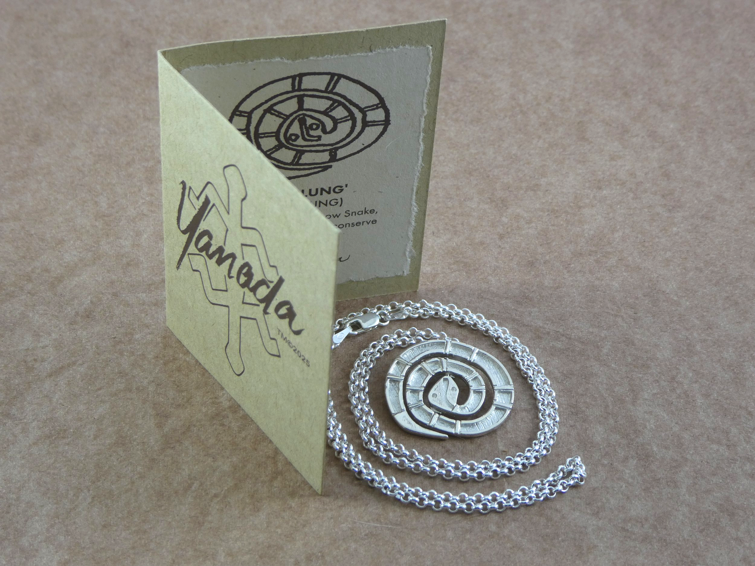 A silver pendant necklace with a maze-like design, accompanied by a charm with the same maze design, displayed on a beige background with a yellow Yamada branded card.
