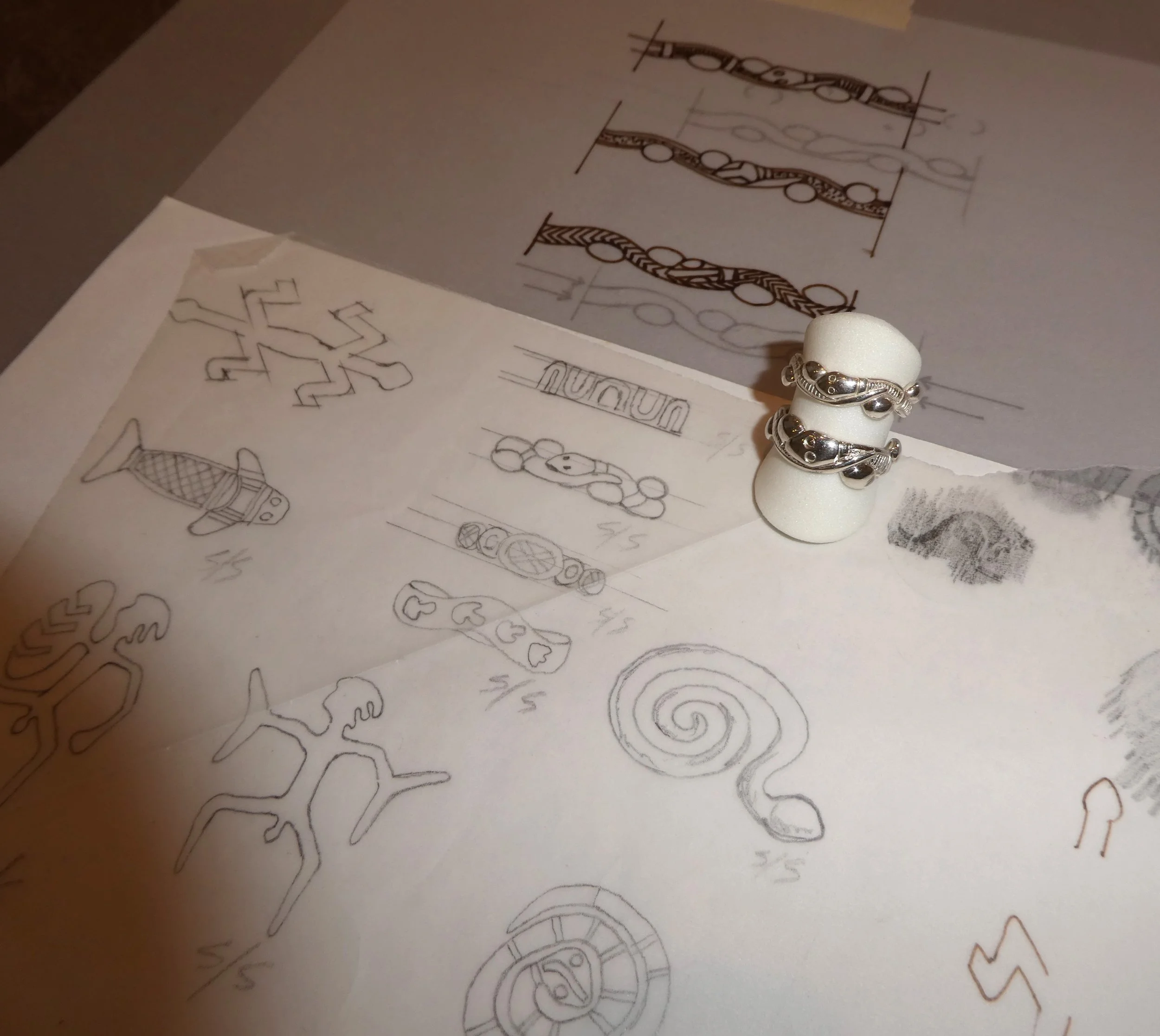 Sketches of jewelry designs on paper, including rings on a small white cylinder, with design sketches in the background.