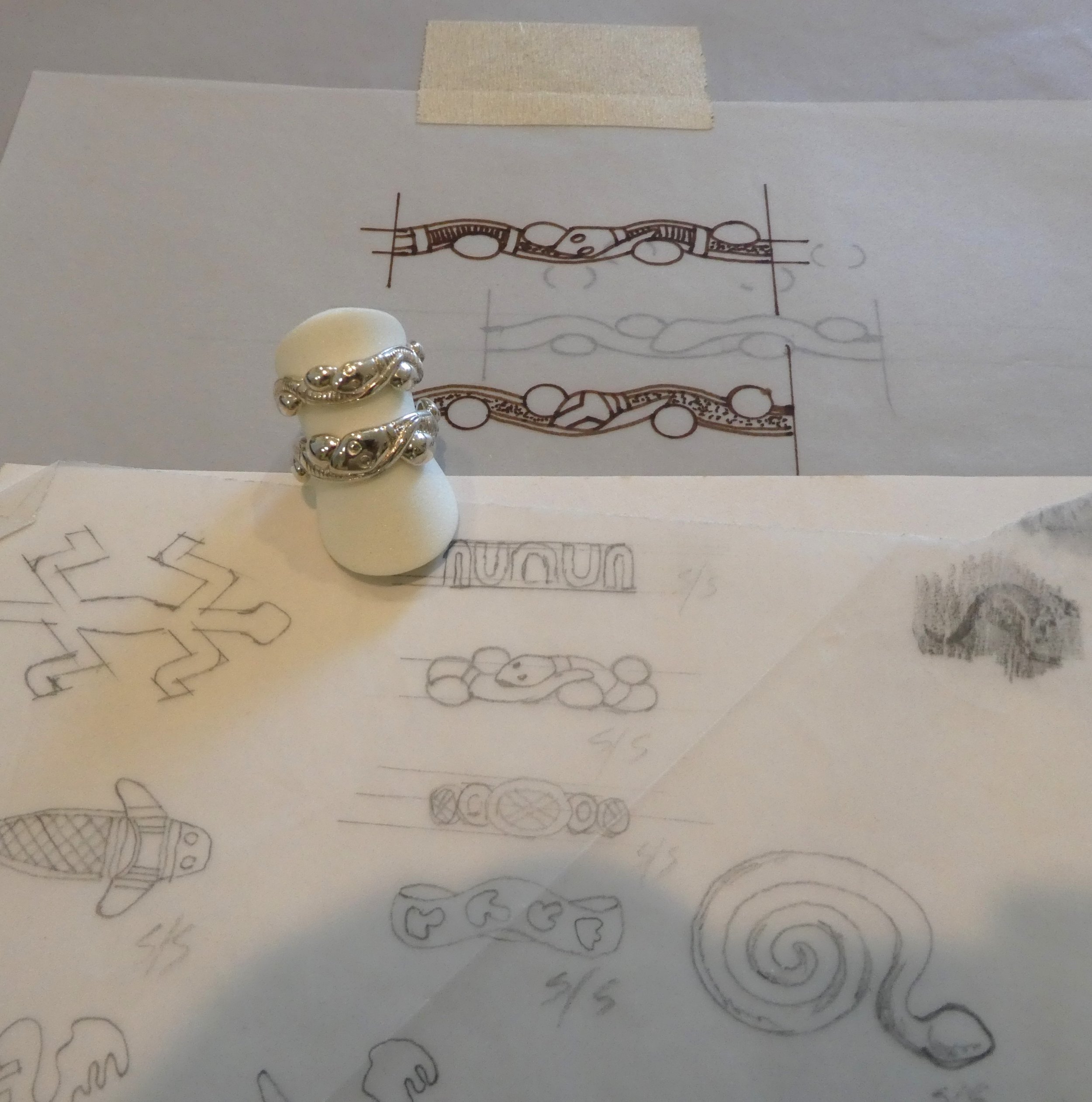 Silver ring with organic, fluid design placed on a white ring holder. The background includes hand-drawn jewelry design sketches on paper.
