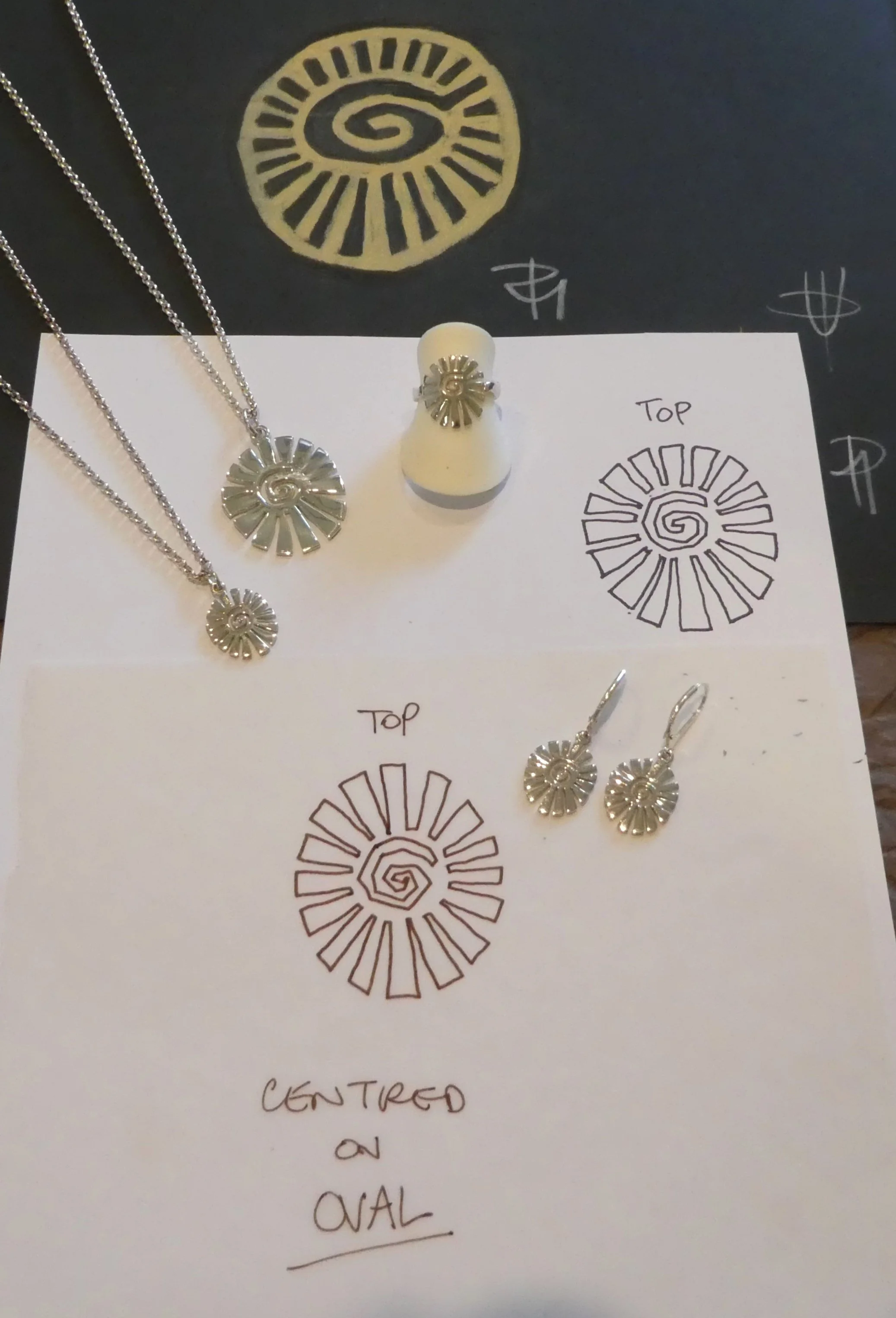 Jewelry set consisting of a necklace, earrings, and a ring, all featuring a spiral sunburst design. The jewelry is displayed on a white paper with hand-drawn and printed diagrams illustrating the design and its different views.