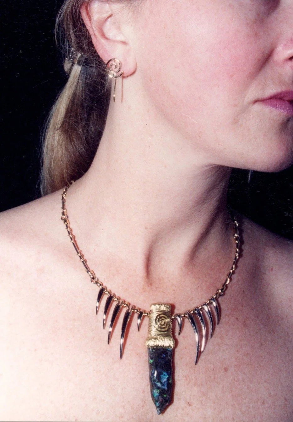 Close-up of a woman wearing a gold and black pendant necklace with spike-shaped charms and an earring with a spiral design, showing her neck and part of her face.