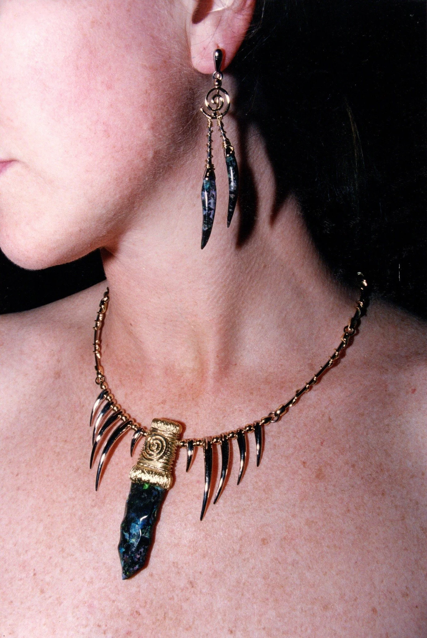 Close-up of a woman wearing a gold and black spiked necklace with a large black stone pendant and matching long earrings with black stones.