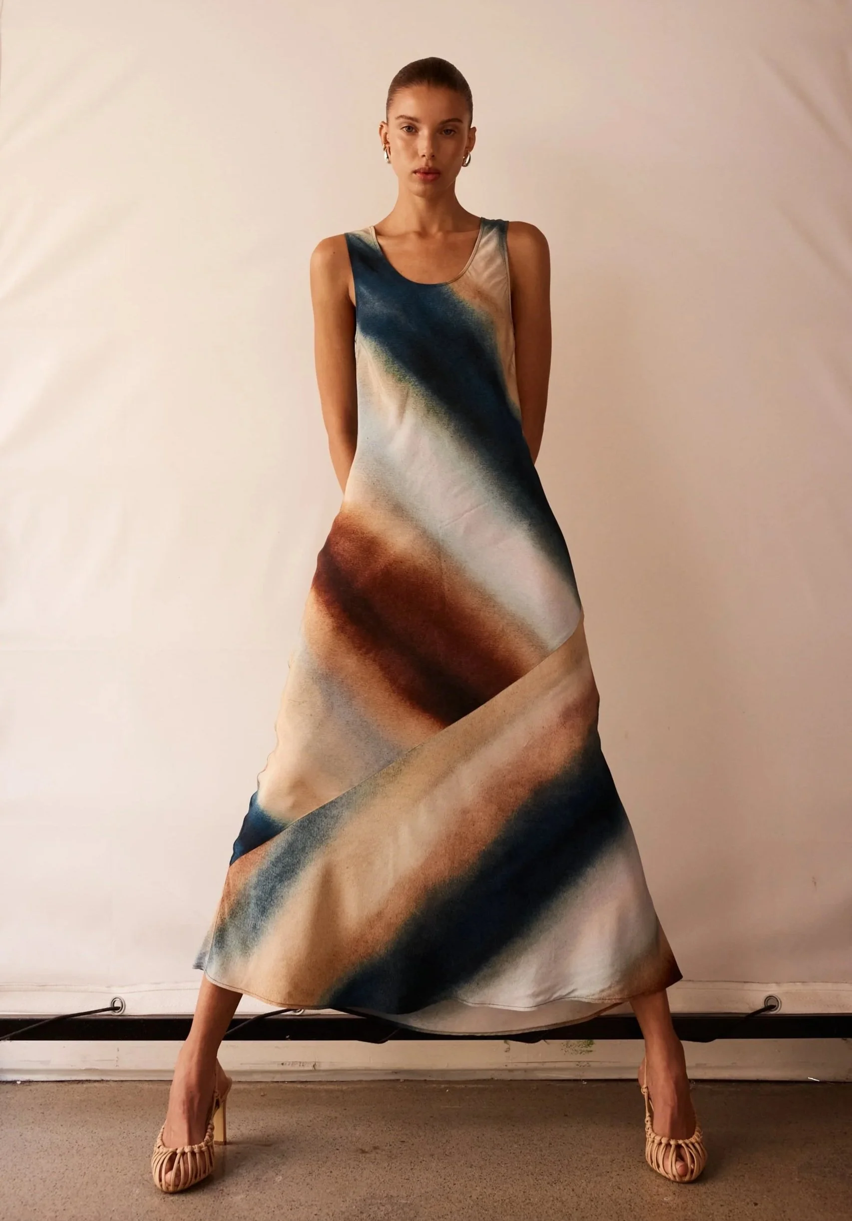 A woman wearing a long, flowing dress with a gradient of swirling colors including shades of blue, brown, cream, and black. She stands with her legs slightly apart and hands behind her back, against a plain light-colored background, wearing beige ope