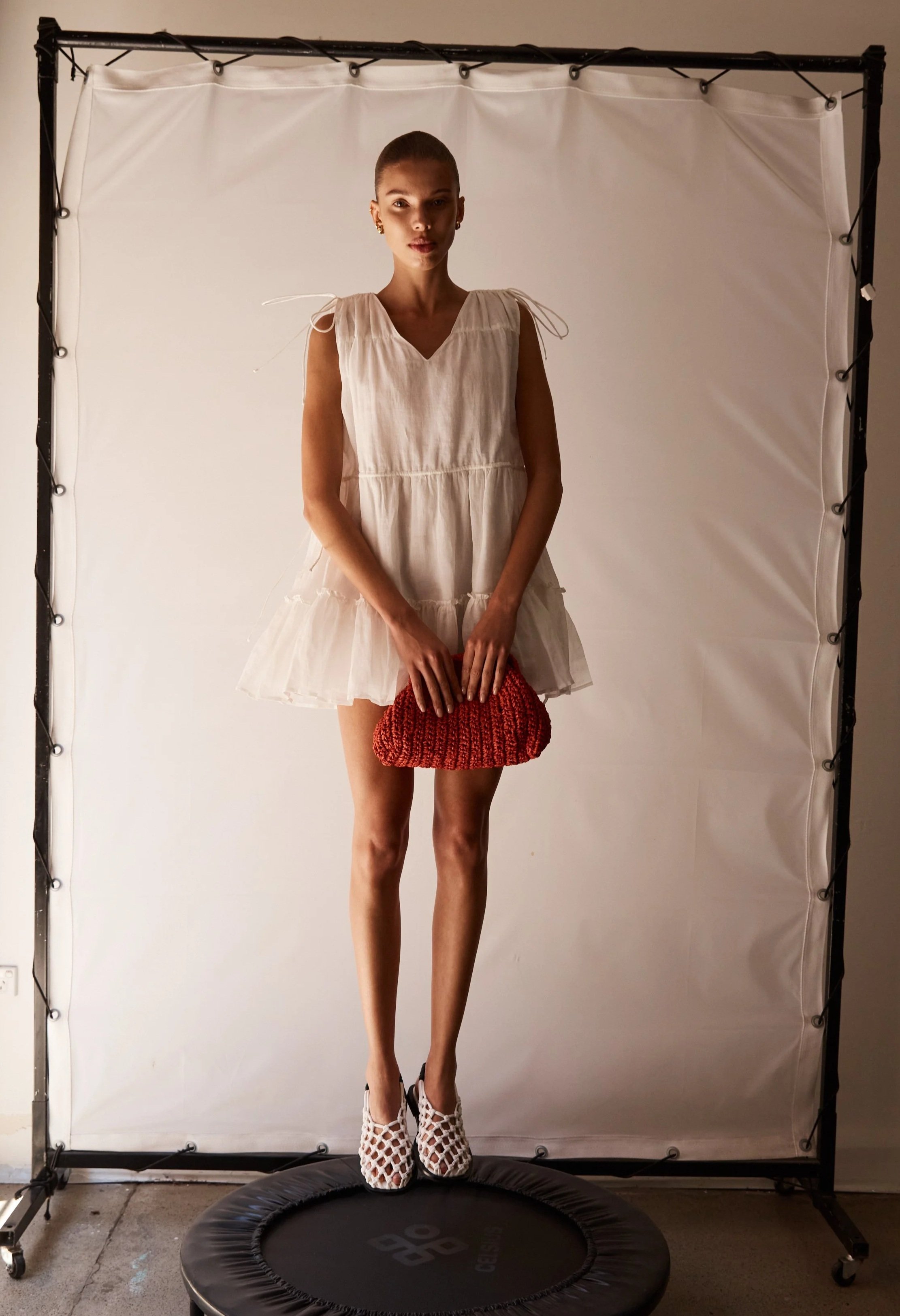 A woman standing on a small trampoline in front of a white backdrops, wearing a sleeveless white dress with a ruffled hem, white patterned open-toe shoes, and holding a knitted red clutch.