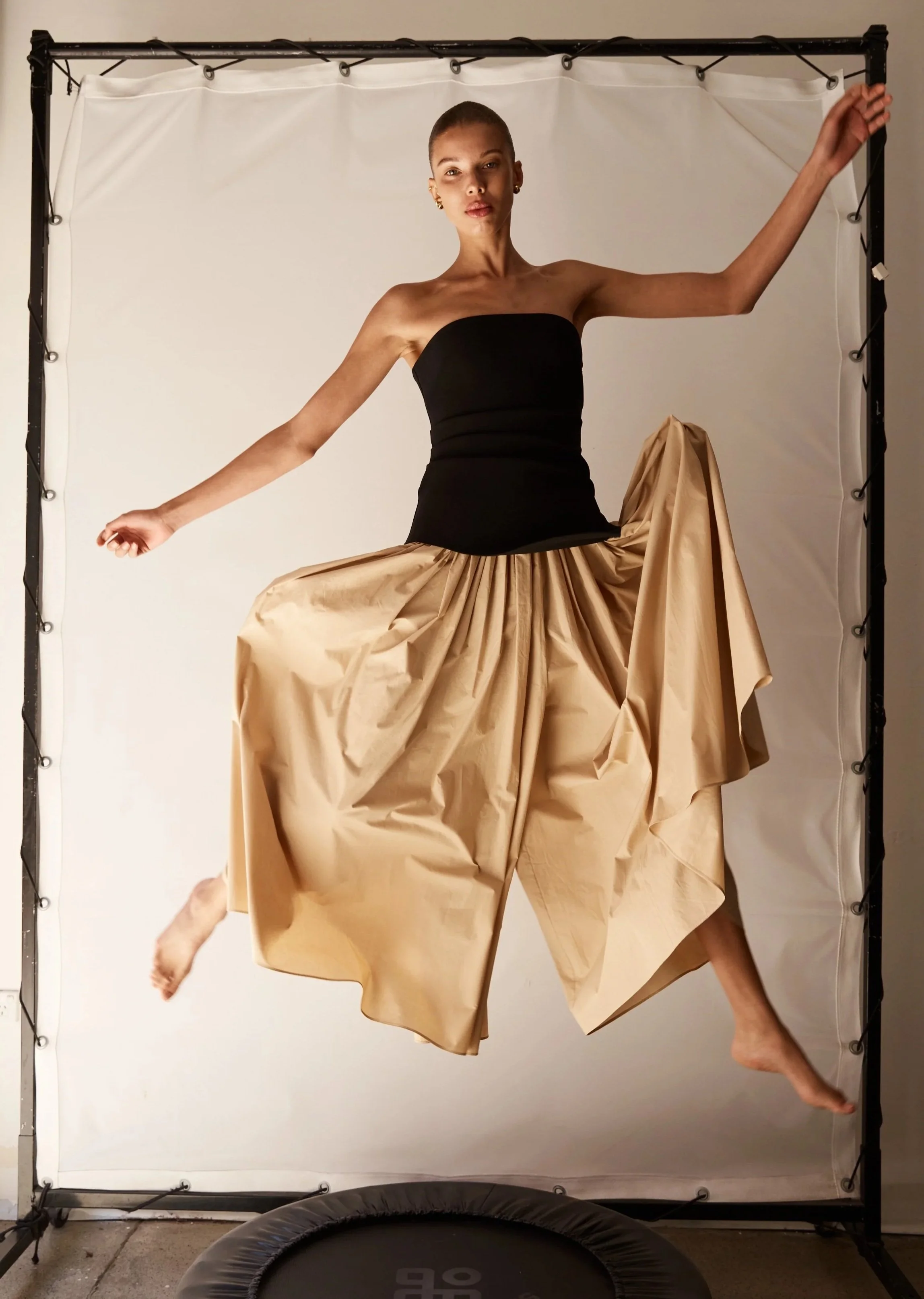 A woman posing with her arms extended, wearing a black strapless top and voluminous beige skirt, floating above a trampoline against a white backdrop.