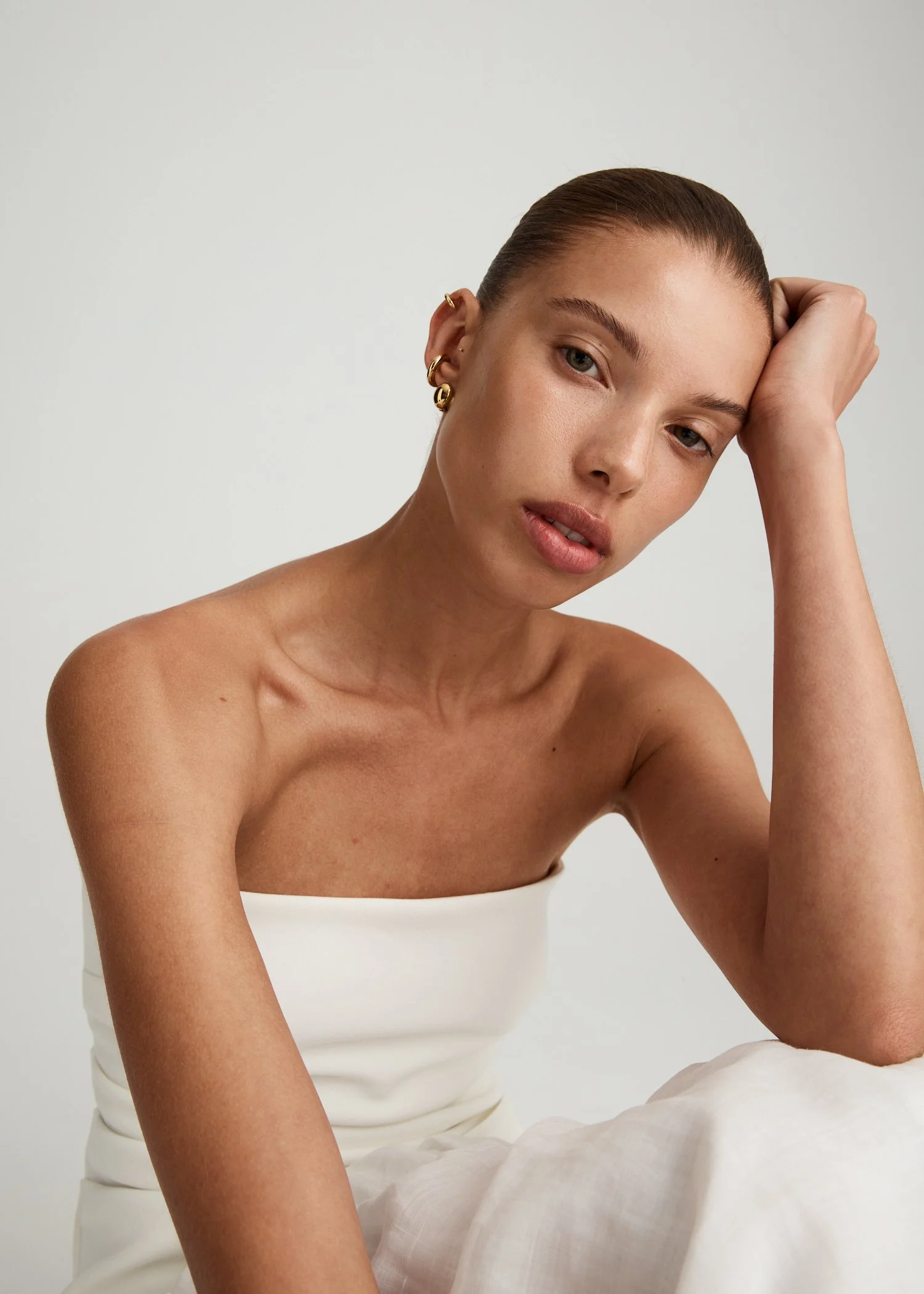 A young woman with fair skin and brown hair styled in a sleek bun, wearing gold earrings, and a white strapless top, sitting against a plain light background.