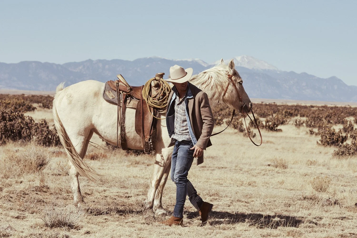 Adam Senn wearing a cowboy hat, jacket, and jeans walking beside a white horse with a saddle and rope in a dry, open landscape with mountains in the background photographed in Colorado. Industrie campaign image