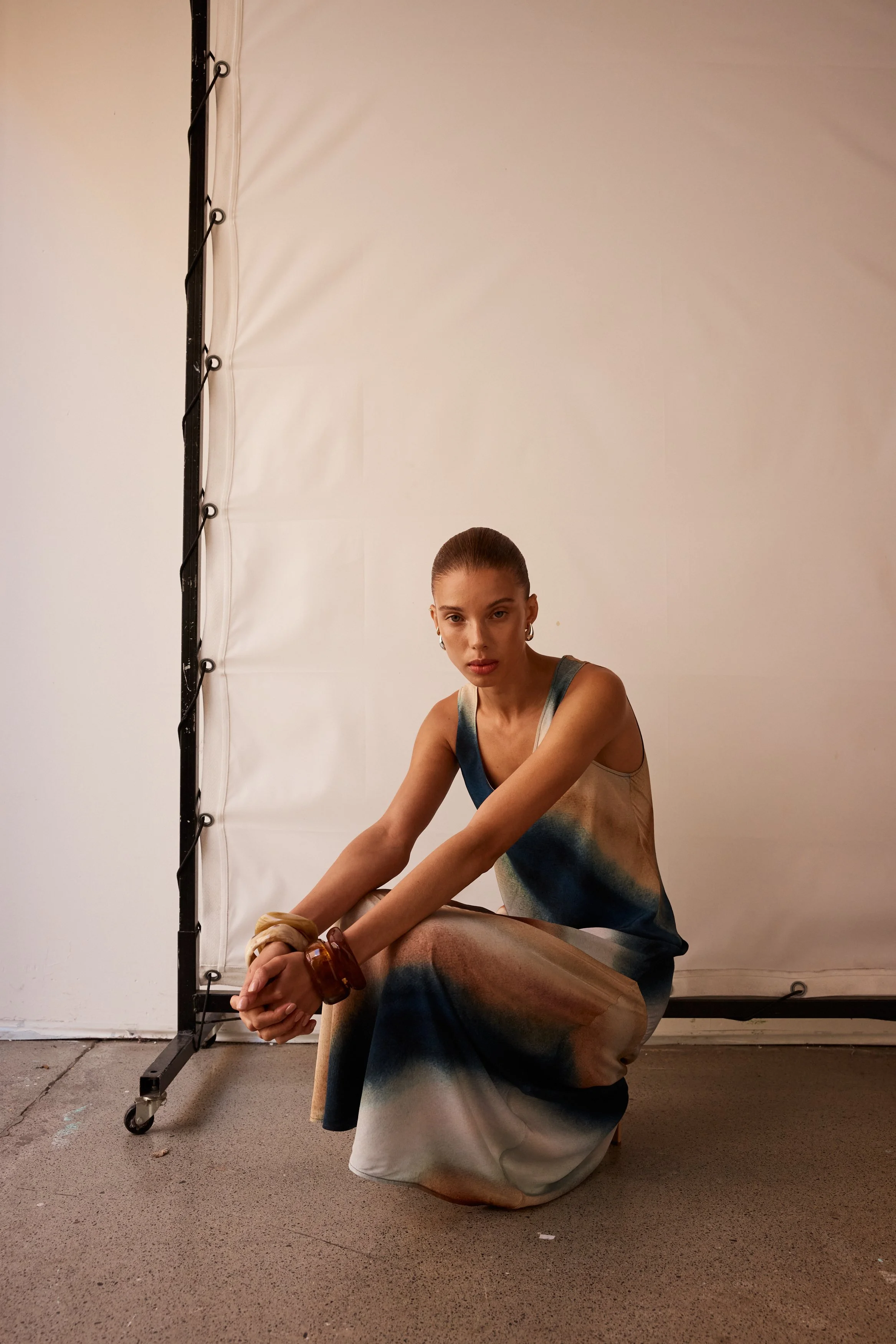 A woman with slicked-back hair crouches on the floor in front of a plain background, wearing a colorful, abstract-print sleeveless dress and large earrings, with her hands clasped together.