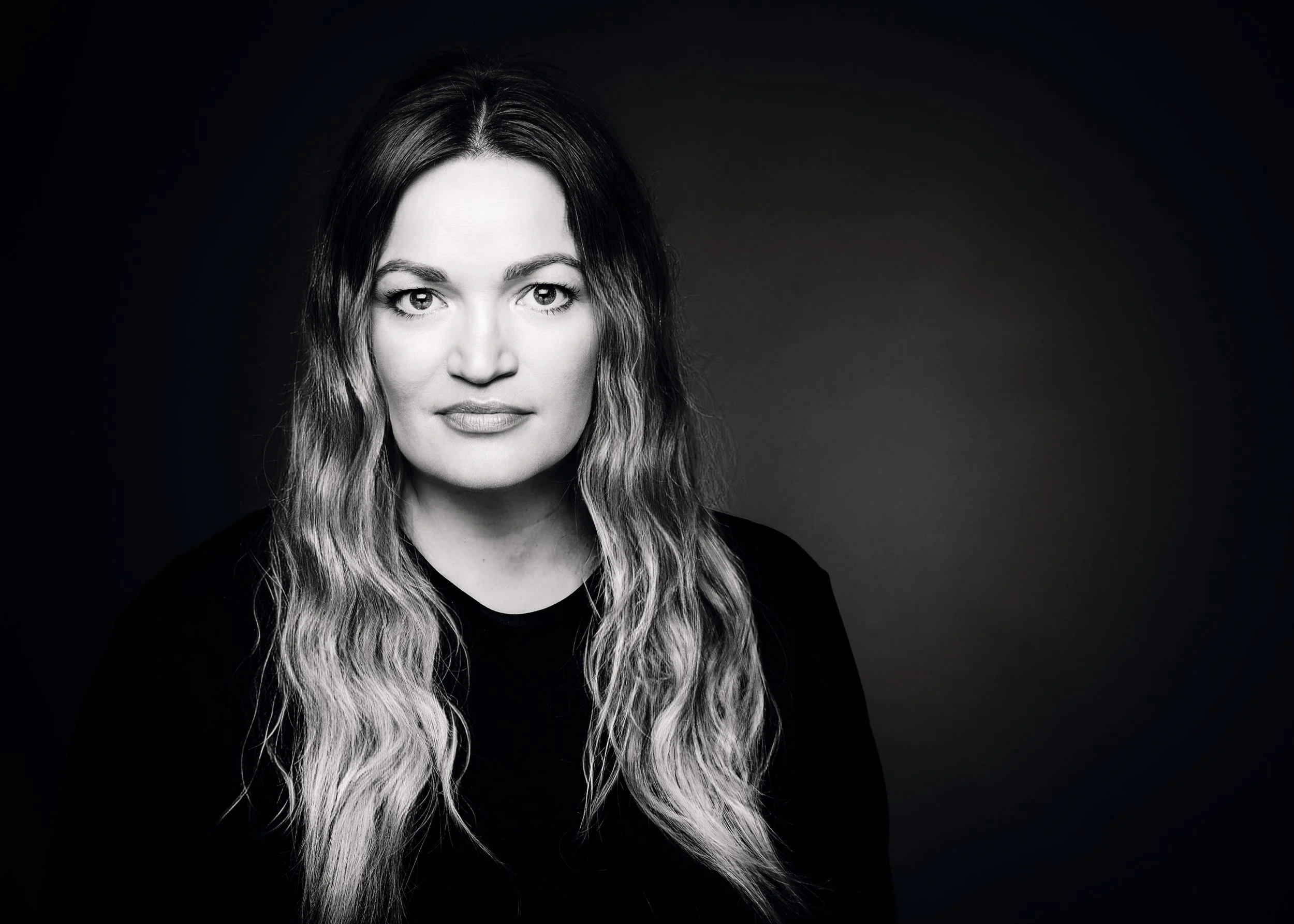 A black and white portrait of a woman with long wavy hair, looking directly at the camera with a neutral expression, on a dark background.