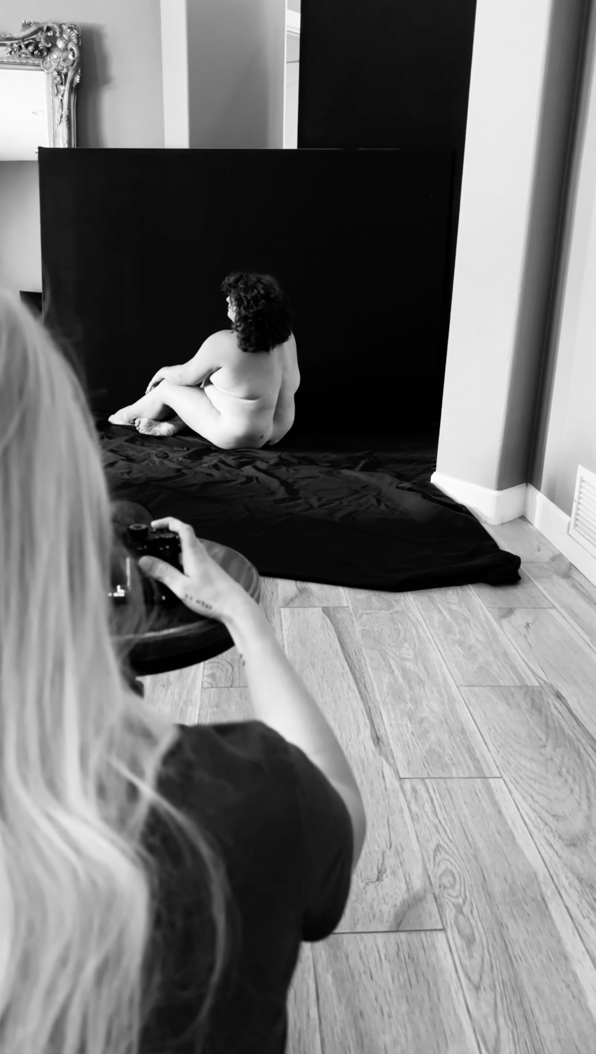 A woman with long blonde hair taking a photo of a naked woman with curly hair sitting on a black backdrop in a room with wooden flooring.
