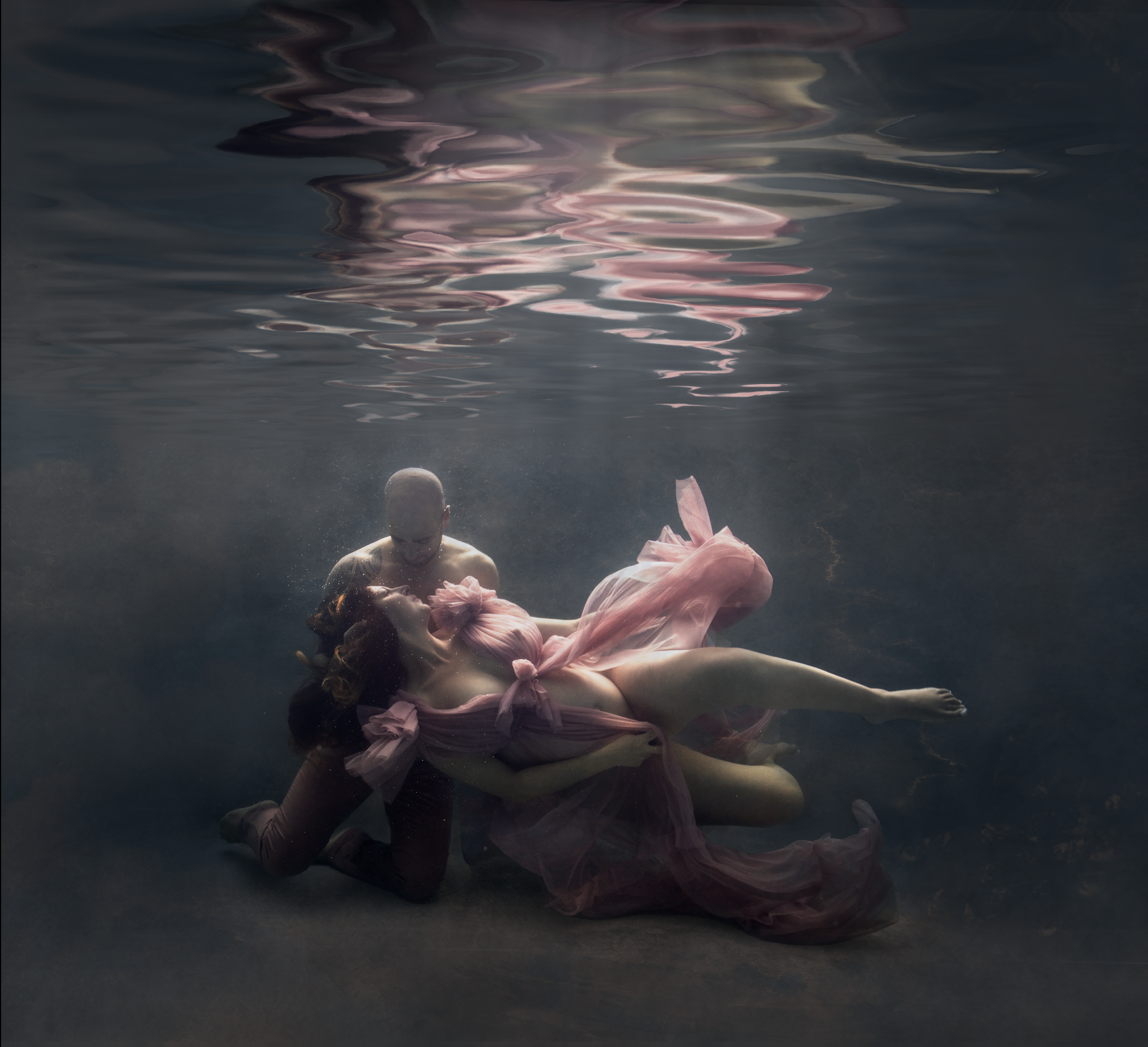 A man and a woman dressed in pink are underwater, with the woman lying on the man's lap, her eyes closed, and the man looking down at her.