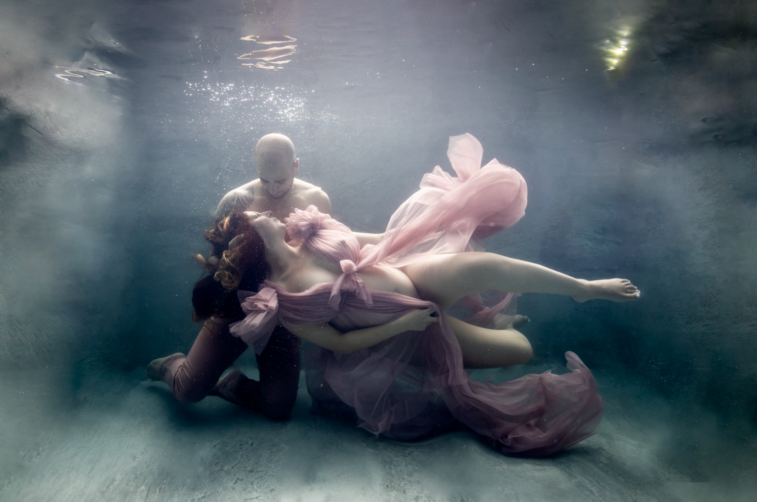 A man and a woman are underwater. The woman is wearing a pink dress and the man is shirtless. The woman is lying back on the man, who is supporting her, with both of them looking at each other.