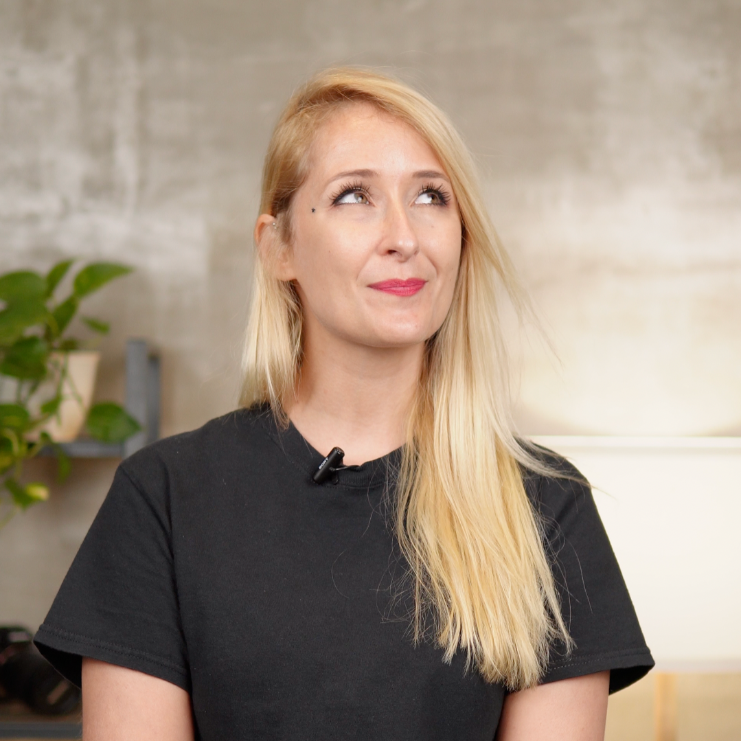 A young woman with long blonde hair, wearing a black t-shirt, looking upward with a smile in an indoor setting.