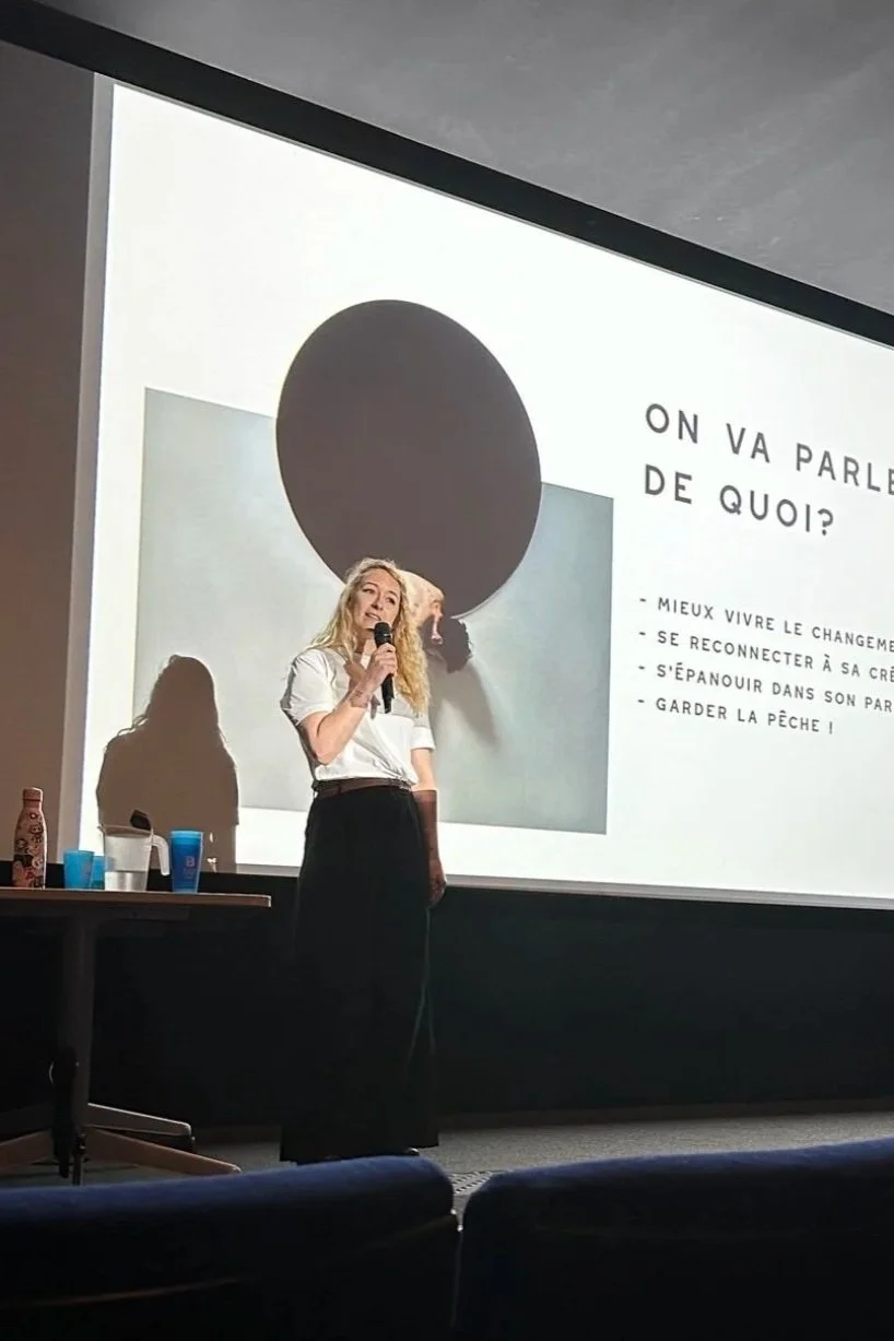A woman is standing in front of a projector screen giving a presentation. She is holding a microphone, has long blond hair, and is dressed in a white top and black skirt. There are some cups and bottles on a table to her left.