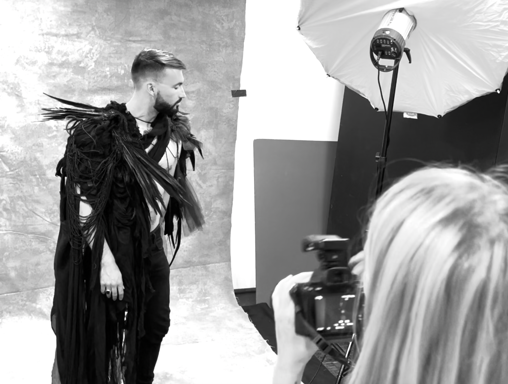 A man stands in front of a photography backdrop, dressed in an elaborate costume made of black fringes and feathers, posing during a photoshoot in a studio with a large umbrella light.
